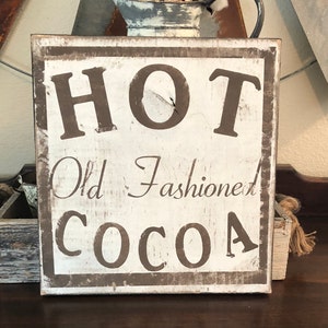 Puede incluir: Un letrero de madera cuadrado y r&uacute;stico con las palabras "HOT Old Fashioned COCOA" en letras marrones. El letrero tiene un aspecto envejecido, con fondo blanco y borde marr&oacute;n, evocando un ambiente acogedor.