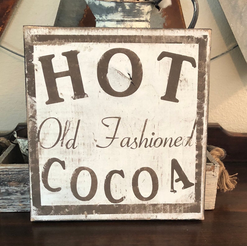 Puede incluir: Un letrero de madera cuadrado y r&uacute;stico con las palabras "HOT Old Fashioned COCOA" en letras marrones. El letrero tiene un aspecto envejecido, con fondo blanco y borde marr&oacute;n, evocando un ambiente acogedor.