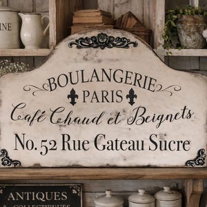 May include: A distressed, cream-colored wooden sign with black lettering that reads "BOULANGERIE PARIS Café Chaud et Beignets No. 52 Rue Gateau Sucre." The sign is decorated with fleur-de-lis and scrollwork. The image evokes a vintage French bakery.