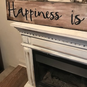May include: A rustic wooden sign with the text "Happiness is" painted in black on a whitewashed background. The sign is sitting on a white fireplace mantel.