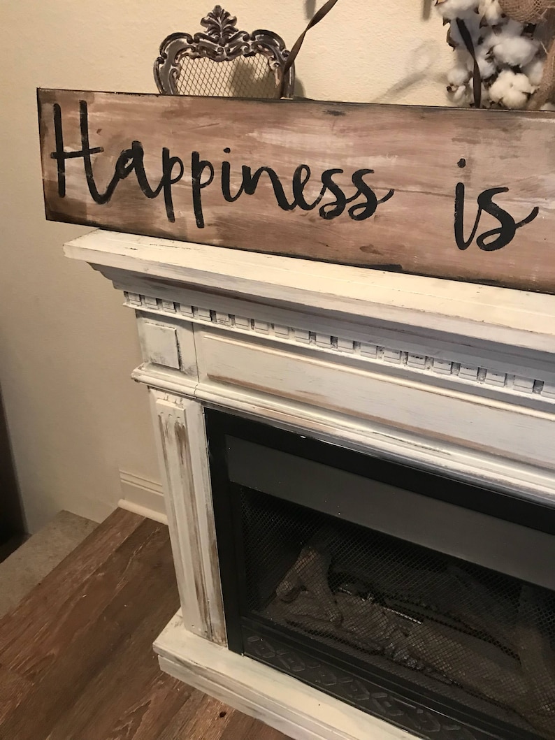 May include: A rustic wooden sign with the text "Happiness is" painted in black on a whitewashed background. The sign is sitting on a white fireplace mantel.