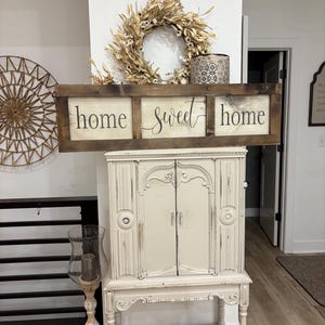 May include: A rectangular wooden wall sign with a dark brown frame and three white panels. The text "home sweet home" is printed in black across the panels. The sign is positioned above a cream-colored cabinet with two doors and ornate legs.