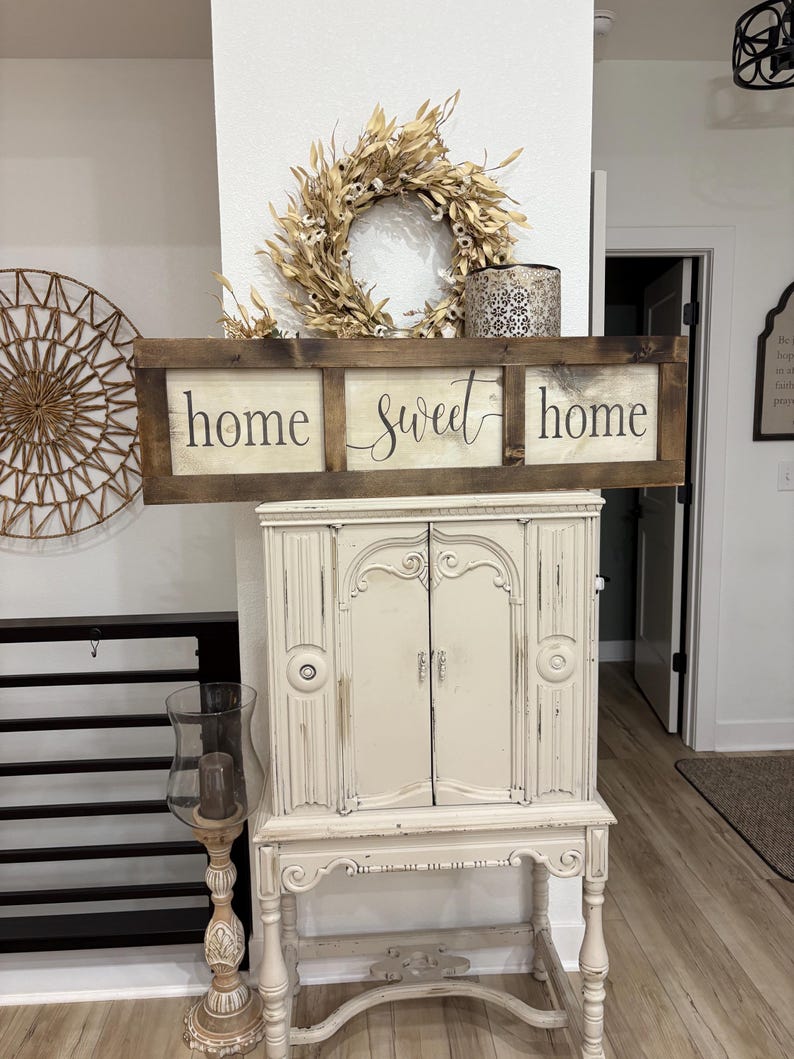 May include: A rectangular wooden wall sign with a dark brown frame and three white panels. The text "home sweet home" is printed in black across the panels. The sign is positioned above a cream-colored cabinet with two doors and ornate legs.