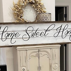 May include: A rectangular wooden sign with the words "Home Sweet Home" in elegant script. The sign has a white background with a distressed wood look and a dark brown frame. A decorative wreath and candle holder are above the sign.