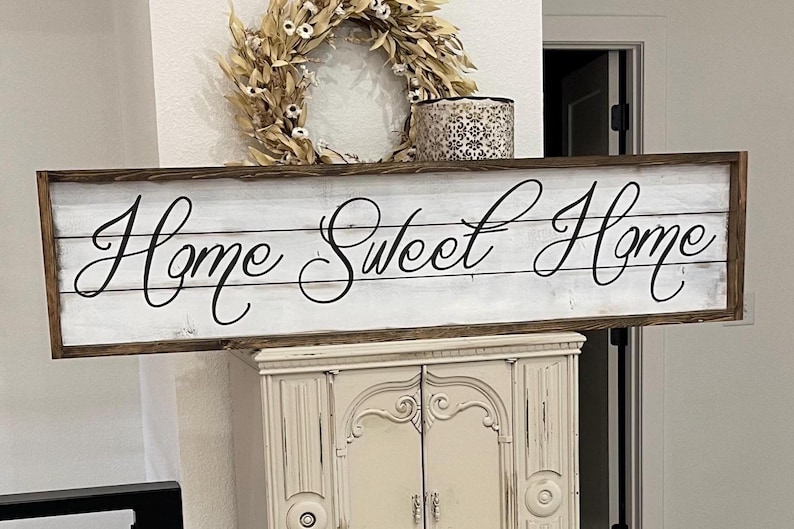 May include: A rectangular wooden sign with the words "Home Sweet Home" in elegant script. The sign has a white background with a distressed wood look and a dark brown frame. A decorative wreath and candle holder are above the sign.