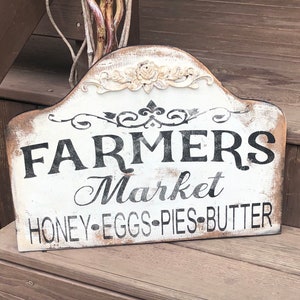 Op de afbeelding: Een wit houten bord met zwarte tekst die "Farmers Market Honey-Eggs-Pies-Butter" luidt. Het bord heeft een verweerde uitstraling en is versierd met een bloemmotief.