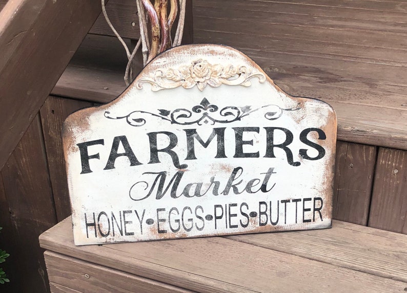 Op de afbeelding: Een wit houten bord met zwarte tekst die "Farmers Market Honey-Eggs-Pies-Butter" luidt. Het bord heeft een verweerde uitstraling en is versierd met een bloemmotief.