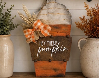 May include: A decorative wooden sign shaped like a coffee cup with whipped cream, featuring the text "HEY THERE pumpkin" in white script. The sign has an orange base, a dark brown middle section, and is adorned with a burlap and orange plaid bow.