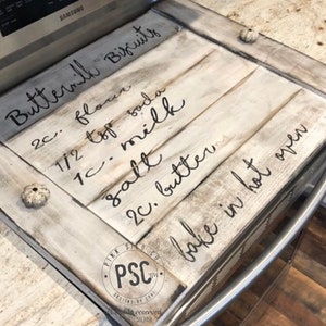 Może przedstawiać: Biała drewniana osłona na płytę grzewczą z czarnym ręcznie pisanym tekstem, kt&oacute;ry brzmi "Buttermilk Biscuits 2 c. mąki 1/2 łyżeczki sody 1 c. mleka s&oacute;l 2 c. masła piecz w gorącym piekarniku". Osłona ma postarzony wygląd i jest ozdobiona małymi muszelkami.