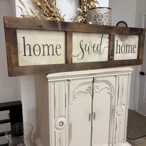 May include: A rectangular wooden sign with a dark brown stained frame and three light-colored panels. The sign features the black text "home sweet home" printed across the panels in a combination of serif and cursive script fonts.