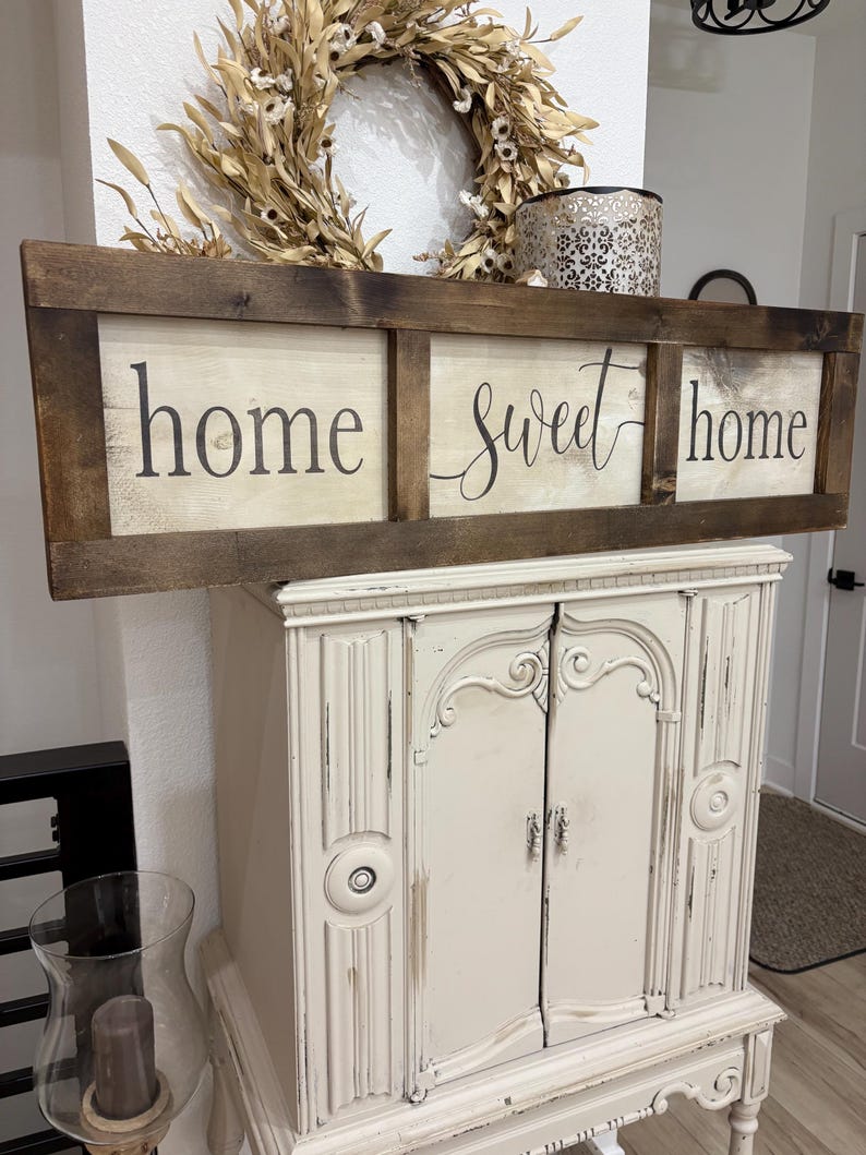 May include: A rectangular wooden sign with a dark brown stained frame and three light-colored panels. The sign features the black text "home sweet home" printed across the panels in a combination of serif and cursive script fonts.
