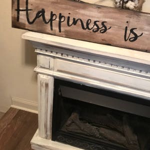 May include: A white painted wooden sign with black lettering that reads "Happiness is". The sign is hanging above a white fireplace with a black grate and faux logs.