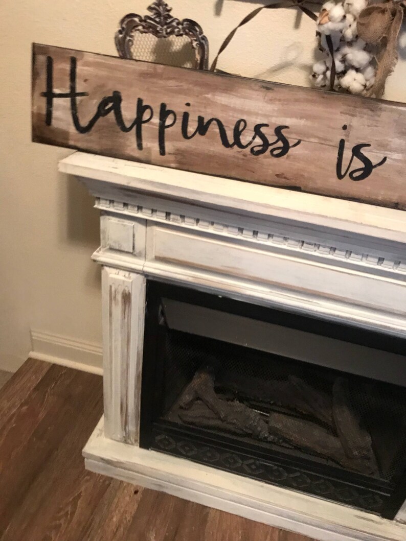 May include: A white painted wooden sign with black lettering that reads "Happiness is". The sign is hanging above a white fireplace with a black grate and faux logs.