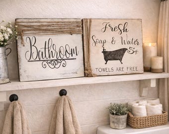 May include: Two square wooden signs with distressed white paint and black lettering. One sign reads Bathroom with decorative scrollwork. The other reads Fresh Soap & Water 5¢ Towels Are Free with a black bathtub graphic. Both feature jute twine accents.