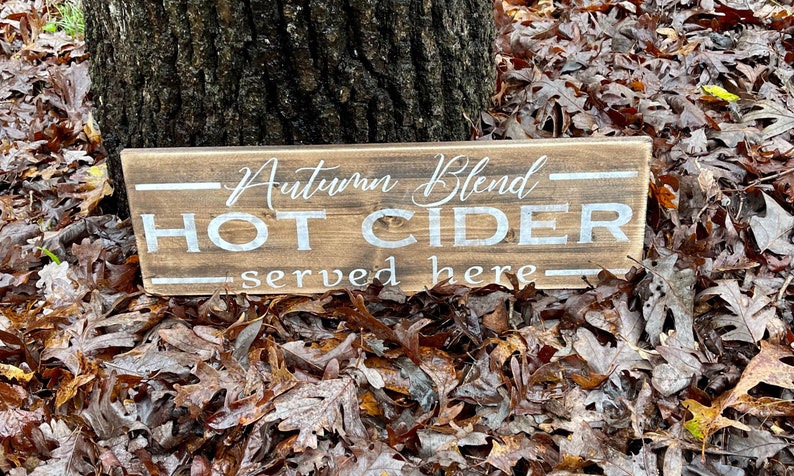 May include: A rectangular wooden sign with the words "Autumn Blend HOT CIDER served here" in white lettering. The sign is set against a backdrop of fallen autumn leaves and a tree trunk.