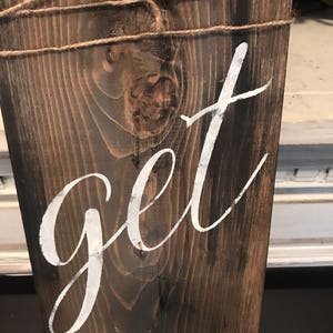 May include: A wooden sign with the words "get in" painted in white on a dark brown background. The sign is tied with twine.