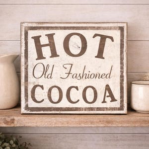Puede incluir: Un letrero r&uacute;stico de madera con las palabras "HOT Old Fashioned COCOA" en letras marrones. Una jarra y una taza de color crema est&aacute;n sobre una estanter&iacute;a de madera, con una manta crema a la derecha.