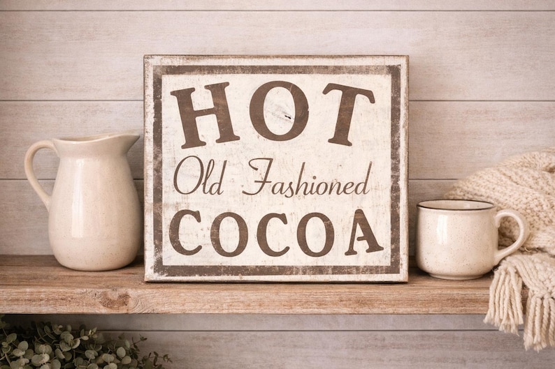 Puede incluir: Un letrero r&uacute;stico de madera con las palabras "HOT Old Fashioned COCOA" en letras marrones. Una jarra y una taza de color crema est&aacute;n sobre una estanter&iacute;a de madera, con una manta crema a la derecha.