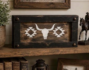 Op de afbeelding: Een rustieke houten kist met een zwart frame en metalen accenten. De kist heeft een witte stierschedel en geometrische ontwerpen op een verweerde houten achtergrond. Western-thema decor.