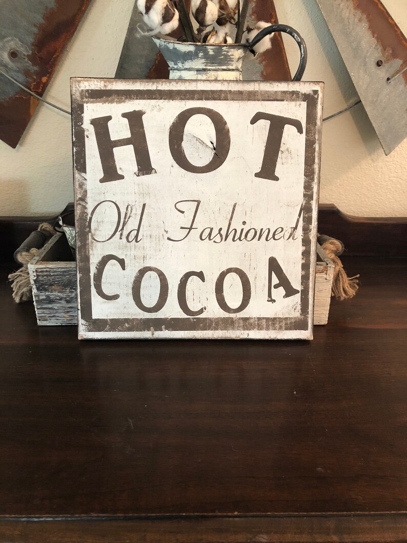 Puede incluir: Un letrero de madera r&uacute;stico con las palabras "HOT Old Fashioned COCOA" en letras marr&oacute;n oscuro. El letrero tiene un fondo blanco y un borde marr&oacute;n oscuro, lo que le da un aspecto vintage. Se muestra sobre una superficie de madera oscura.