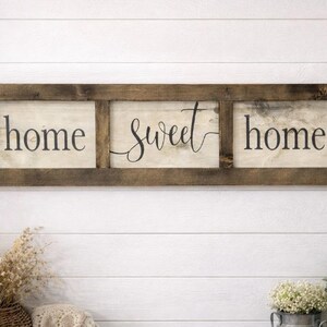 May include: A rectangular wooden wall sign featuring three separate panels framed in dark brown wood. The off-white panels display the black text "home sweet home" in a combination of serif and cursive script fonts, arranged horizontally.