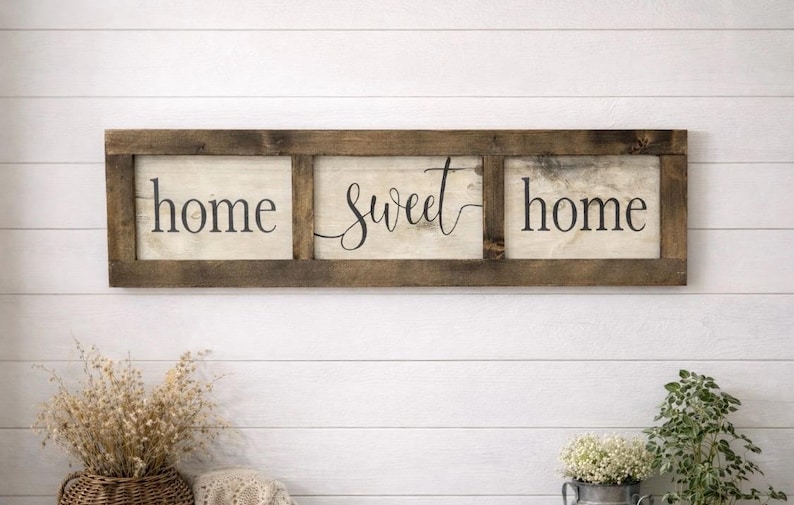 May include: A rectangular wooden wall sign featuring three separate panels framed in dark brown wood. The off-white panels display the black text "home sweet home" in a combination of serif and cursive script fonts, arranged horizontally.