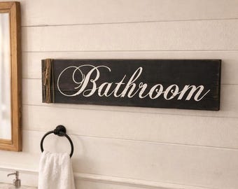May include: A rustic black wooden sign with the word "Bathroom" in elegant white script. The sign is mounted on a white shiplap wall, with a towel ring and a white towel hanging below. A wooden framed mirror is visible on the left.