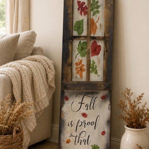May include: A tall, rectangular wooden sign featuring a dark brown frame with decorative metal corner accents. The upper section resembles a window pane with painted autumn leaves in red, orange, and green. The lower section displays the text Fall is proof that change is Beautiful in black script, surrounded by small painted leaves.