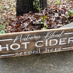 May include: A rectangular wooden sign with the words "Autumn Blend HOT CIDER served here" in white lettering. The sign is made of dark brown wood and is placed outdoors.