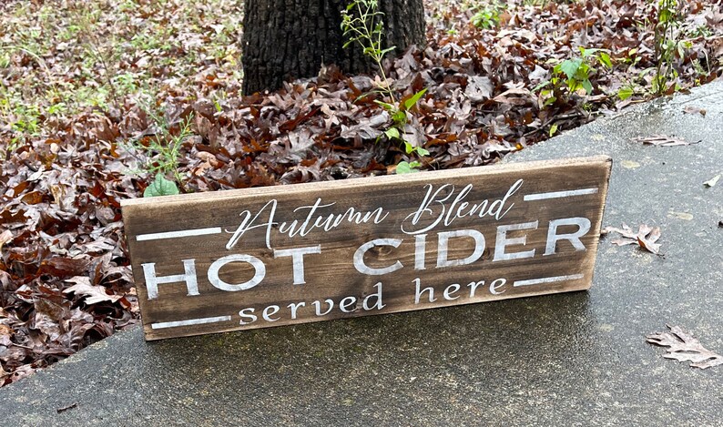 May include: A rectangular wooden sign with the words "Autumn Blend HOT CIDER served here" in white lettering. The sign is made of dark brown wood and is placed outdoors.