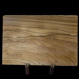 May include: A rectangular, sandstone-like serving board with a natural, layered pattern in shades of brown and tan. The board is supported by two small, cylindrical wooden legs, creating a raised presentation. The edges have a slightly rough, natural finish.