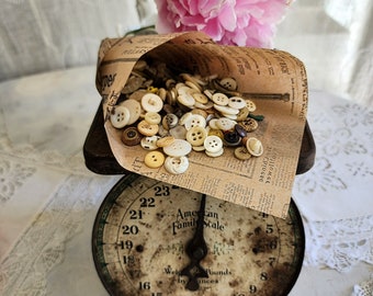 May include: A collection of assorted small round buttons in various shades of white, cream, and brown, contained within a folded piece of vintage-style printed paper, resting on a weathered metal American Family Scale with a circular dial.
