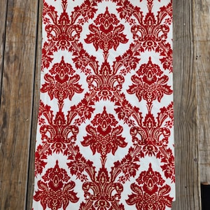 May include: A length of fabric displaying a repeating red damask pattern against a white backdrop. The elaborate design incorporates floral and scroll motifs. The material is suitable for home decor or upholstery.