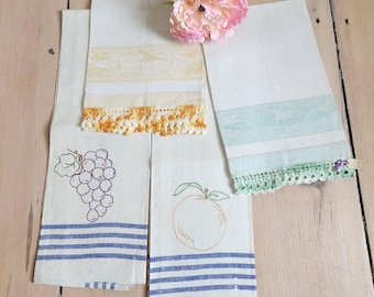 Puede incluir: Tres paños de cocina rectangulares blancos con diseños de frutas bordados, incluyendo un racimo de uvas moradas y una naranja. Los paños tienen bordes de rayas azules y adornos de ganchillo en amarillo y verde, con una flor rosa encima.