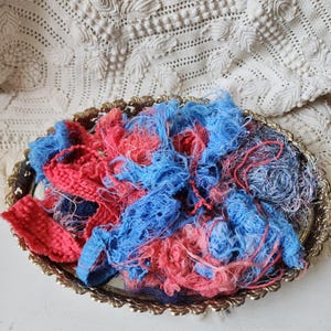 May include: A collection of frayed, textured fabric strips in vibrant shades of red, blue, and dark navy, piled together on an oval-shaped, ornate gold-toned metal tray with a scalloped edge and a reflective mirrored base.