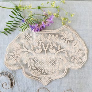 May include: A beige, intricate, hand-crocheted lace doily featuring a delicate, open-work grid pattern with scalloped edges. The doily is positioned beneath a sprig of small, light purple flowers and green leaves on a light gray fabric surface.
