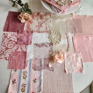 May include: A collection of pink and cream fabric swatches with various textures and patterns, including floral prints, stripes, and lace. A crocheted doily and a silk ribbon add to the vintage aesthetic. A rose and greenery are placed on the top left.
