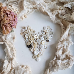 May include: A heart shape made of white buttons on a white background with white lace surrounding the heart.