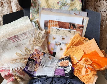 May include: A collection of assorted craft supplies inside a black box, including yellow floral fabric, orange lace, grey textured fabric, vintage paper ephemera with bee motifs, and various textile scraps with intricate embroidery and crochet details.
