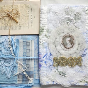 May include: A quilted fabric cover featuring a floral pattern, a circular lace doily, a vintage-style portrait medallion, and gold trim. Beside it, a stack of blue fabric scraps, lace, and a page from the Atlantic Monthly with lace and beads.