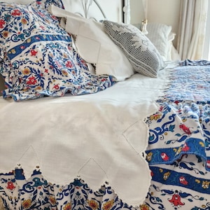 Könnte beinhalten: Ein Bett mit weißem Kopfteil und Bettwäsche. Die Bettwäsche umfasst eine weiße Bettdecke mit Muschelmuster und Kissen. Die gemusterte Bettdecke und die Kissenbezüge weisen ein blaues, rotes und gelbes Blumenmuster auf. Die Bettdecke hat einen gewellten Rand.