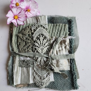 Puede incluir: Un collage textil en capas con varios tonos de verde salvia y oliva, incluyendo arpillera, terciopelo y encaje. Una aplicación central de encaje blanco está atada con una cinta verde salvia, y tres flores cosmos de color rosa y blanco descansan encima.