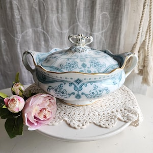 Pode incluir: Uma travessa de servir vintage com tampa, adornada com motivos florais azuis e bordas douradas. Possui duas alças e um adorno decorativo na tampa. Apresentada sobre uma toalha de crochê branca e um pedestal branco. Rosas cor-de-rosa em primeiro plano.