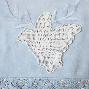May include: A light blue fabric textile featuring a white lace butterfly appliqué centered on the surface. The fabric is decorated with embroidered leaf sprigs above the butterfly and a wide, intricate blue lace border along the bottom edge.