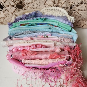 Peut inclure: Une pile de chutes de tissus, de dentelles et de rubans assortis, disposés en dégradé de couleurs allant du violet clair, vert menthe et bleu en haut, vers diverses nuances de rose et corail en bas, agrémentée d'un collier de perles roses.