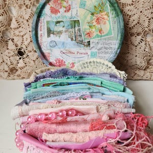 Pode incluir: Uma tampa de lata redonda com um design de postal vintage, motivos florais e texto em italiano, sobre uma coleção de retalhos de tecido, rendas e contas cor-de-rosa translúcidas em camadas, com vários tons de rosa, azul e verde.