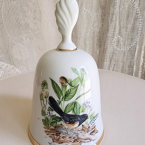 May include: White porcelain bell with a gold trim and a detailed painting of a black and white bird perched on a branch with green leaves and purple flowers. The bell has a white, twisted handle and the text "Roger Tory Peterson" is printed on the bottom.