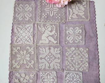 May include: A square lavender fabric mat featuring eleven cream-colored crocheted lace squares arranged in a grid. The lace squares display various intricate geometric and floral patterns, including one square depicting a cherub figure.