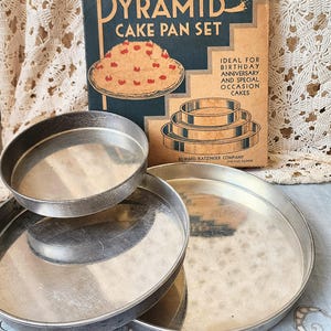 May include: A collection of vintage silver cake tins of different sizes. The image includes a vintage advertisement for a "Pyramid Cake Pan Set". The advertisement features a cake illustration and the text "Ideal for Birthday, Anniversary and Special Occasion Cakes."