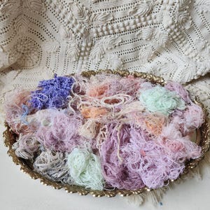Op de afbeelding: Een ovaalvormig, sierlijk dienblad van goudkleurig metaal gevuld met een verzameling verwarde, getextureerde garenresten in diverse pasteltinten zoals lavendel, perzik, mintgroen en zachtroze, naast een kleine pluk donkerpaarse vezels.